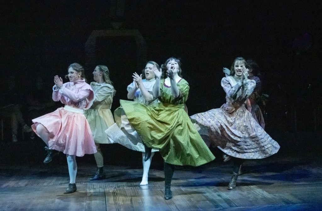 A group of five women in colorful vintage dresses performing a dynamic dance on stage, with a dark background highlighting their movement.