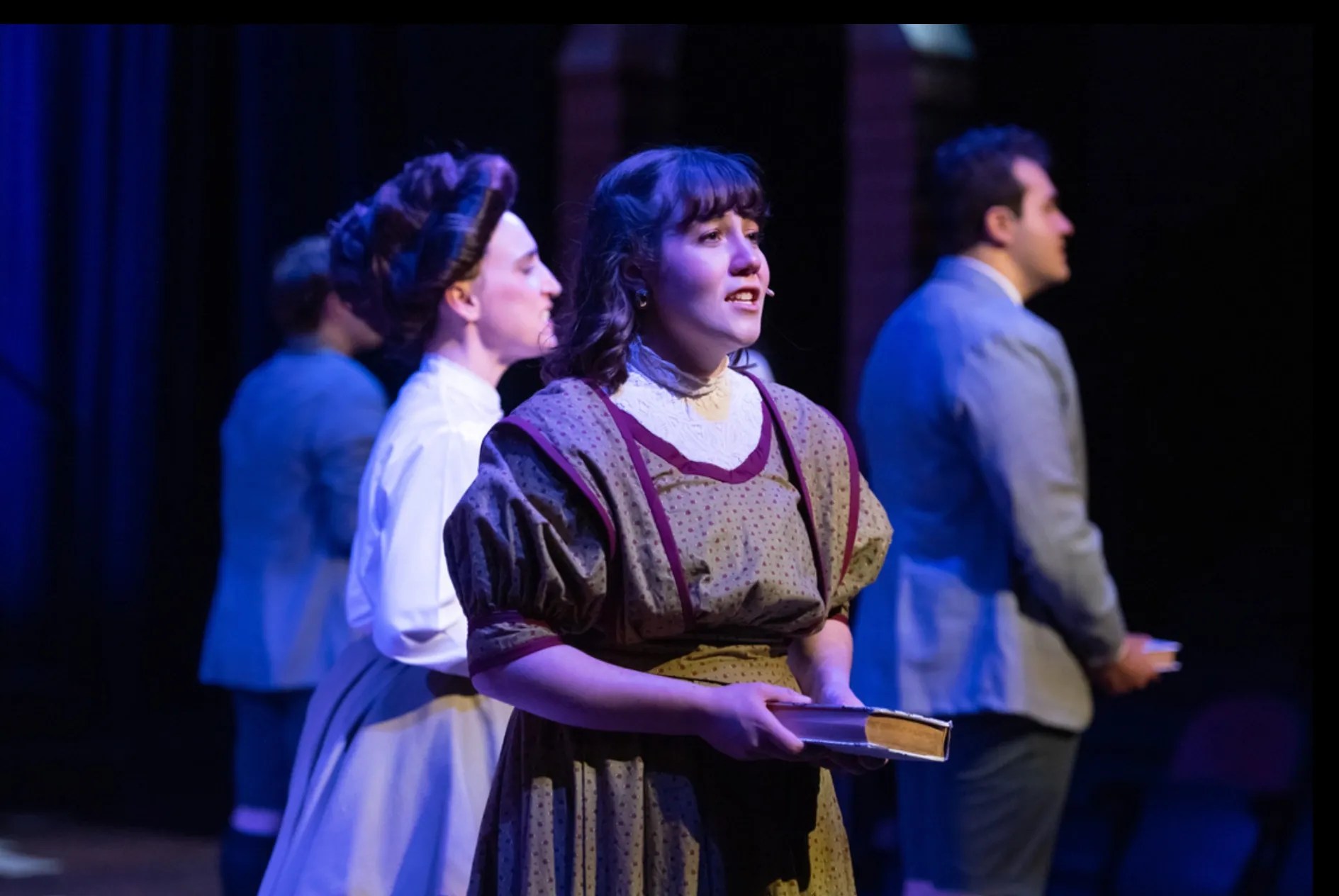 A performer on stage holding a book, wearing a vintage dress, with other cast members in the background, during a theatrical performance.