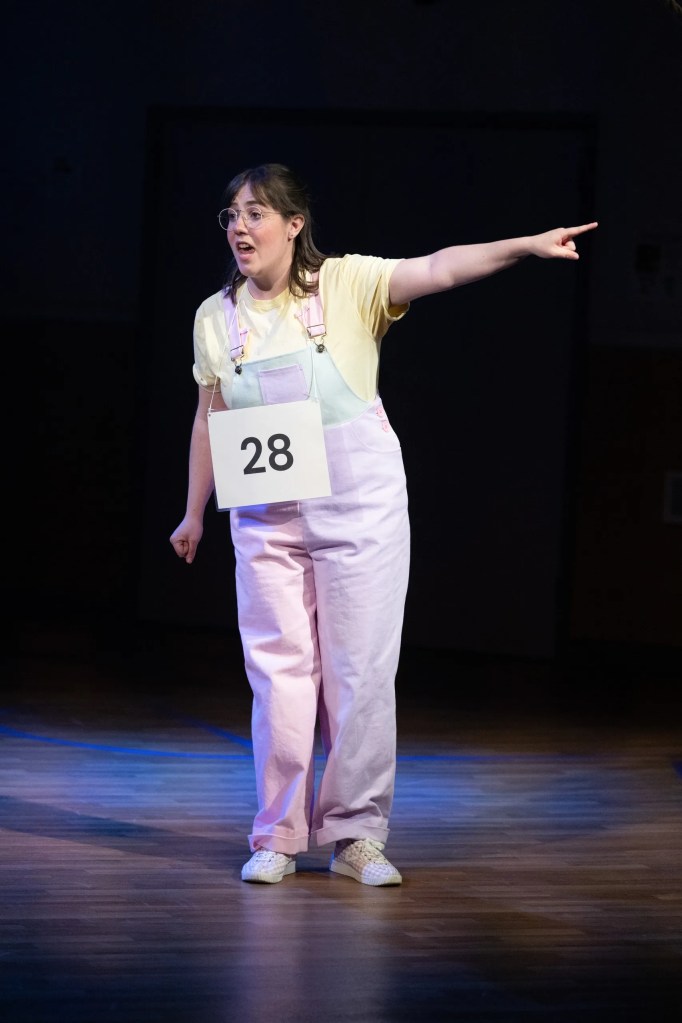 A performer in a colorful outfit holding a sign with the number 28, dramatizing a scene while pointing in a stage setting with focused lighting.