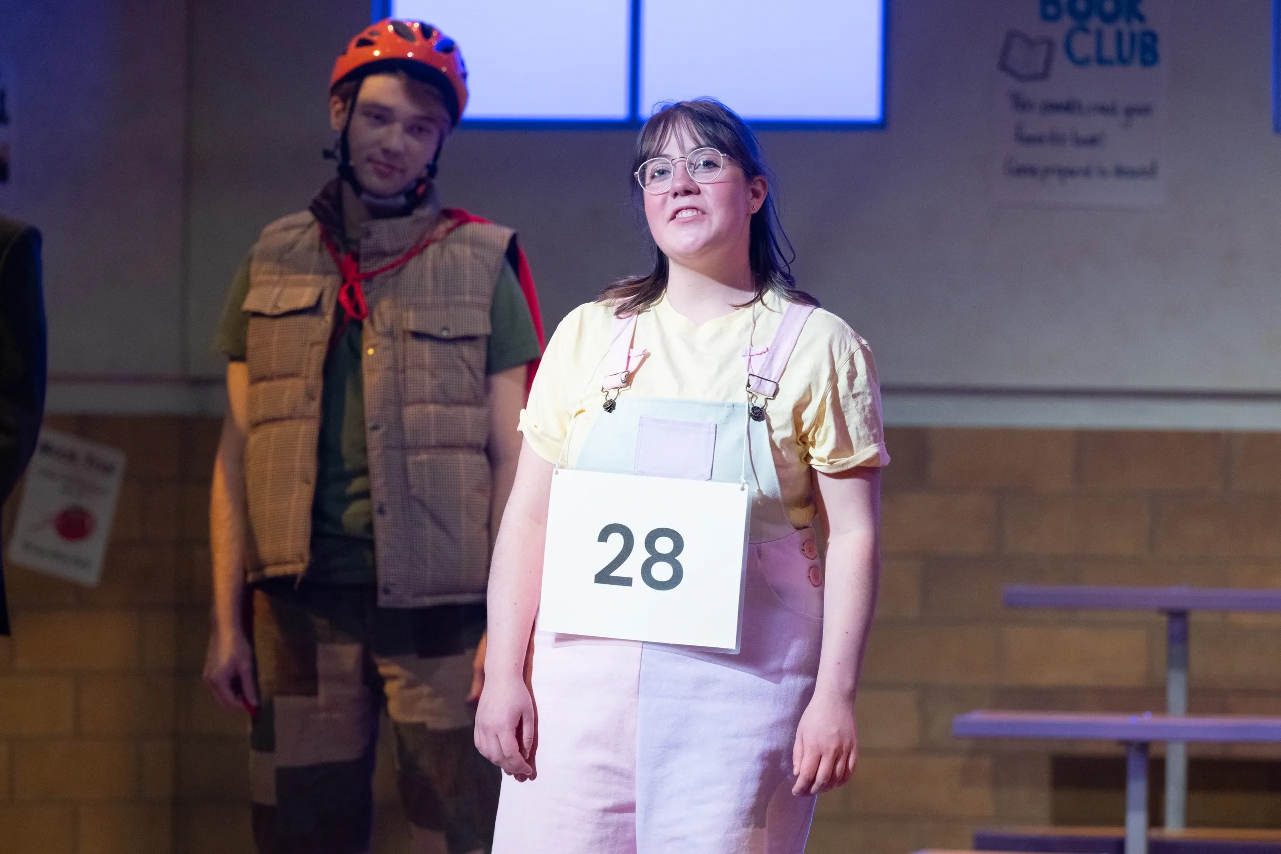 A young woman in a pastel pink outfit with a sign displaying the number 28, standing on stage with a serious expression, while a male figure in the background wears a helmet and vest.
