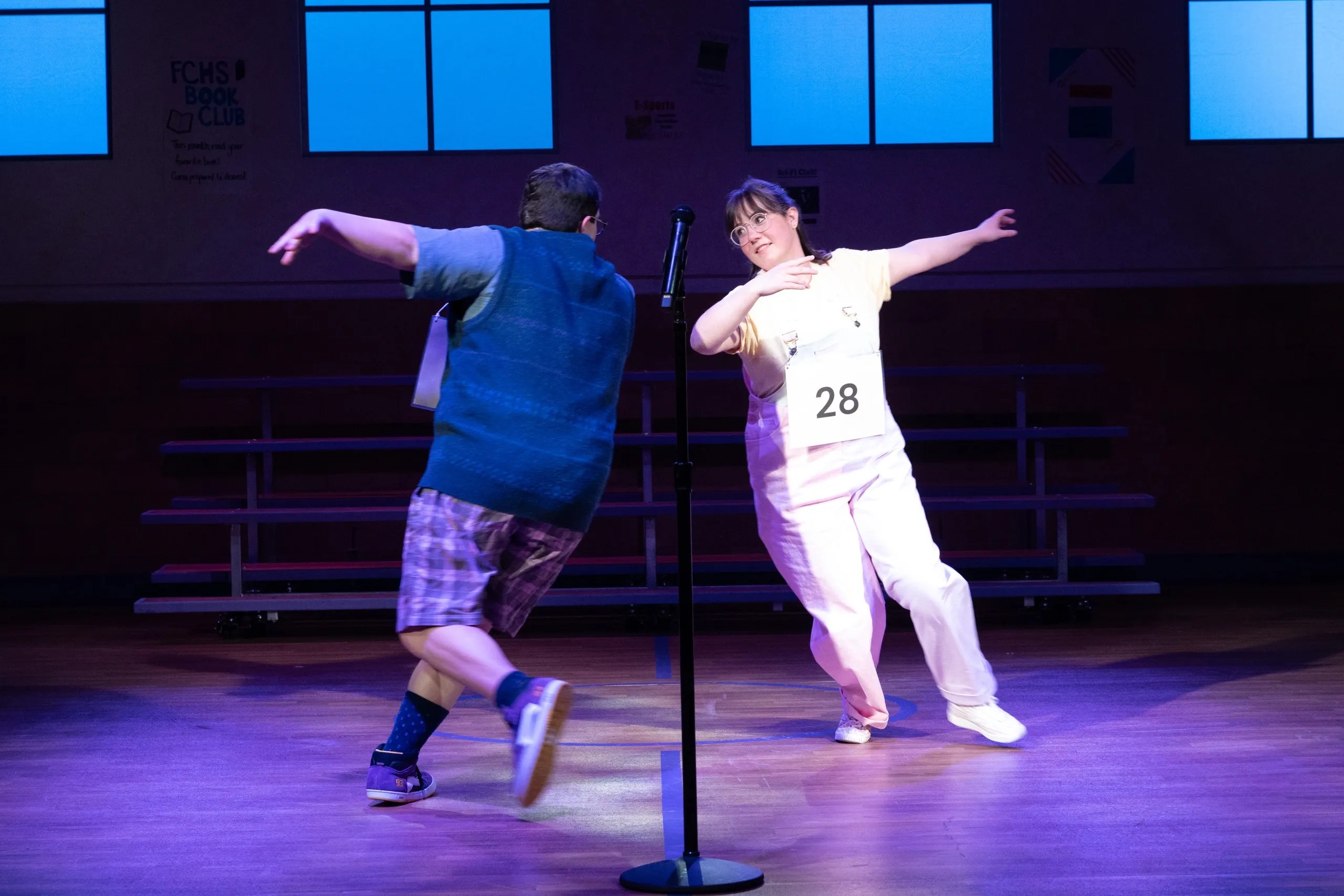 Two performers on stage during a theatrical competition, one wearing a blue sweater and the other in a yellow shirt with a number 28. They appear to be dancing and engaging with the audience.