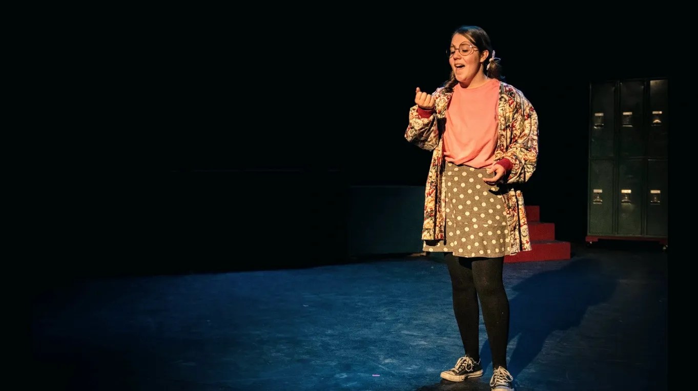 A performer stands on stage with a microphone, wearing a colorful patterned jacket, pink shirt, and polka dot skirt, with a spotlight highlighting her against a dark background.