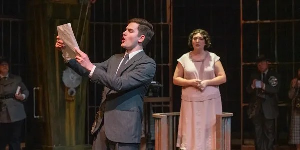 Photographer: John Eisele
A male actor in a suit passionately reads a piece of paper while a female actor stands behind him, wearing a dress and observing. The stage is set with a dark background and multiple actors in costume in the distance, suggesting a dramatic performance.
