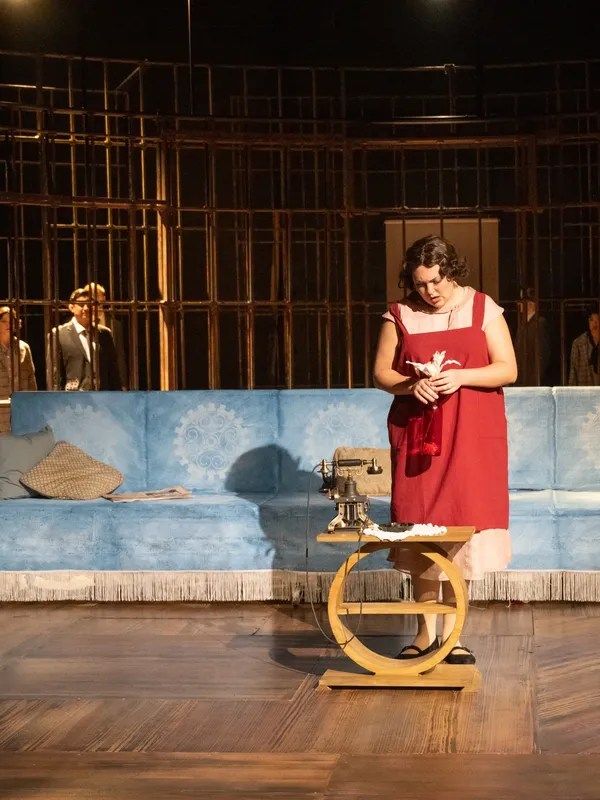 Photographer: John Eisele
A woman in a red dress stands by a sewing machine, appearing contemplative, with a blue couch and wooden backdrop visible in the theater setting.