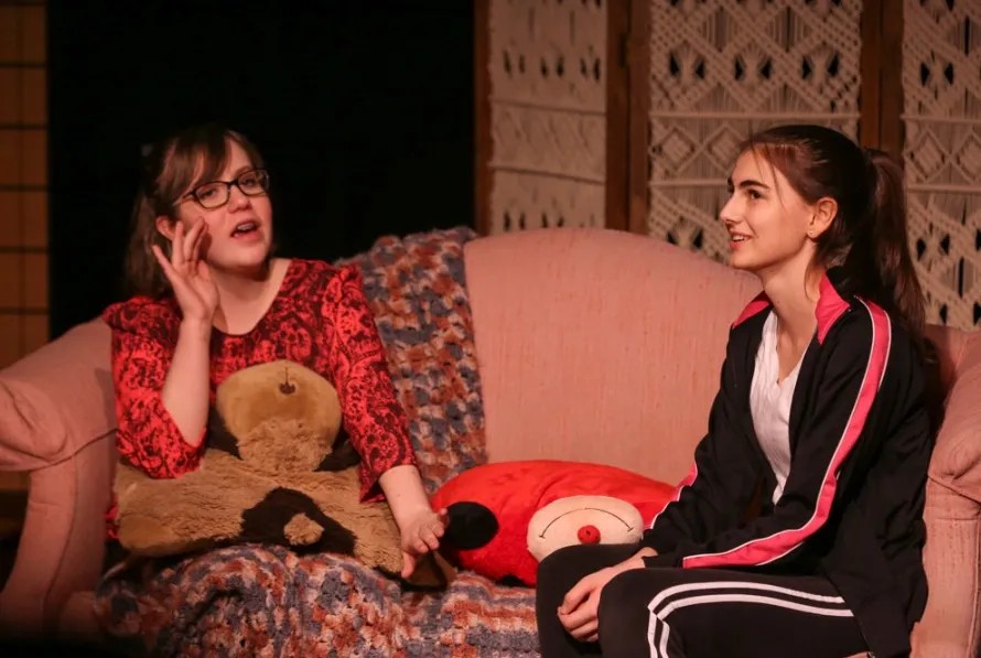 Two actresses performing a scene on stage, sitting on a pink couch. One is holding a teddy bear and gesturing expressively, while the other listens attentively. The background features a decorative screen.